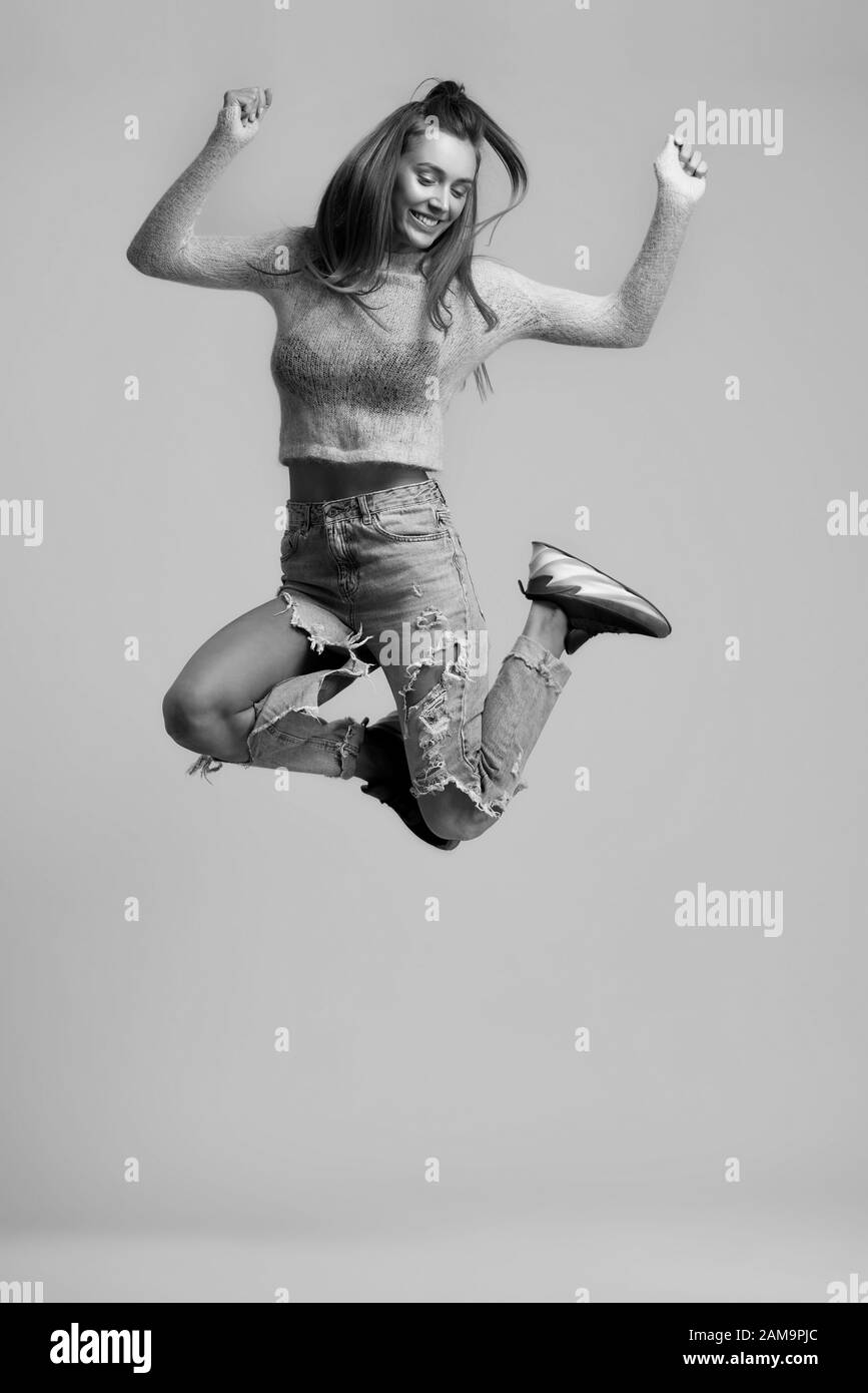 Schwarz-Weiß-Foto von lustigen fröhlichen Mädchen mit langen Haaren, die Beine im Sprung hochheben, mit kurzem Oberteil mit langen Ärmeln, gerippten Jeans, schwarzen Turnschuhen auf isoliertem Hintergrund im Studio. Stockfoto