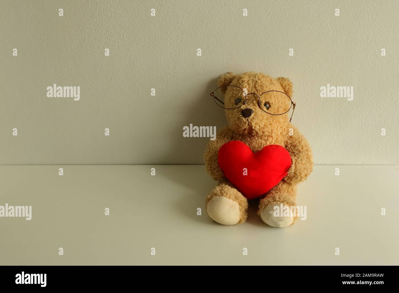 Teddybär-Puppe mit Brillen auf weißem Holztisch, der rotes Herz hält und Kamera, Liebeskonzept betrachtet Stockfoto