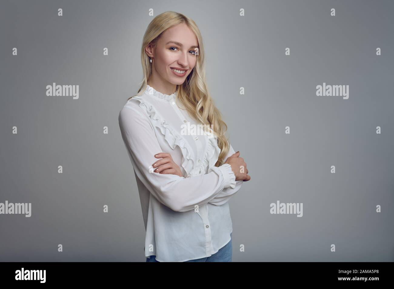 Junge und schöne langhaarige blonde Frau in weißer Bluse, die mit gefalteten Armen steht und mit einem Lächeln auf die Kamera blickt. Stockfoto