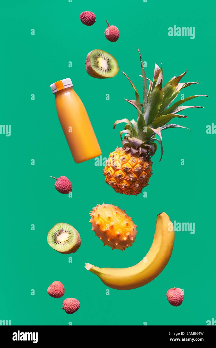 Auswahl an tropischen Früchten und orangefarbenen Smoothie-Flaschen auf grünem Hintergrund. Ananas, Kiwano, Kiwi, Lichee und Banane - exotische Früchte, Levitation Stockfoto