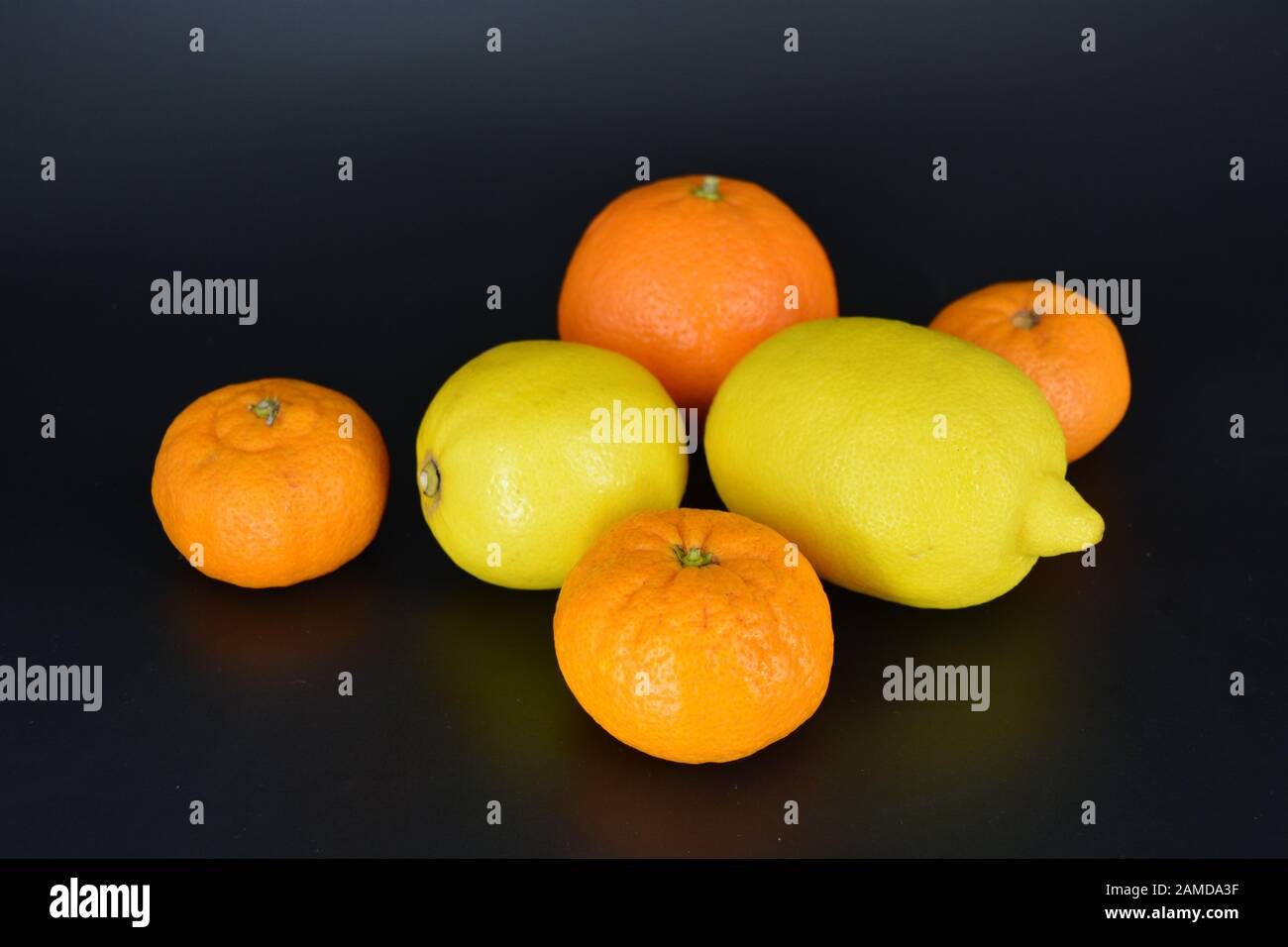 Frische helle Früchte, zwei gelbe Zitronen, drei kleine orangefarbene Mandarinen, eine große Mineralola liegt auf schwarzem Kunststoffhintergrund. Lecker und süß. Stockfoto