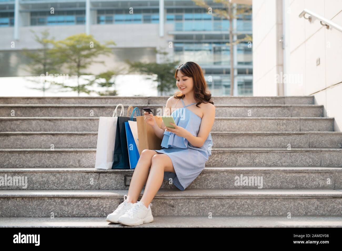 Schöne asiatische Frau mit Einkaufstaschen, die auf Treppen sitzen, während sie auf das Smartphone schaut und die Kreditkarte hält. Stockfoto