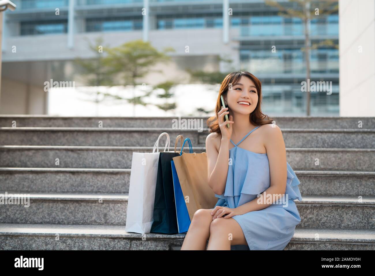 Fröhliche junge Asiatin mit Einkaufstaschen, die auf dem Handy telefonieren und auf einer Treppe sitzen. Stockfoto