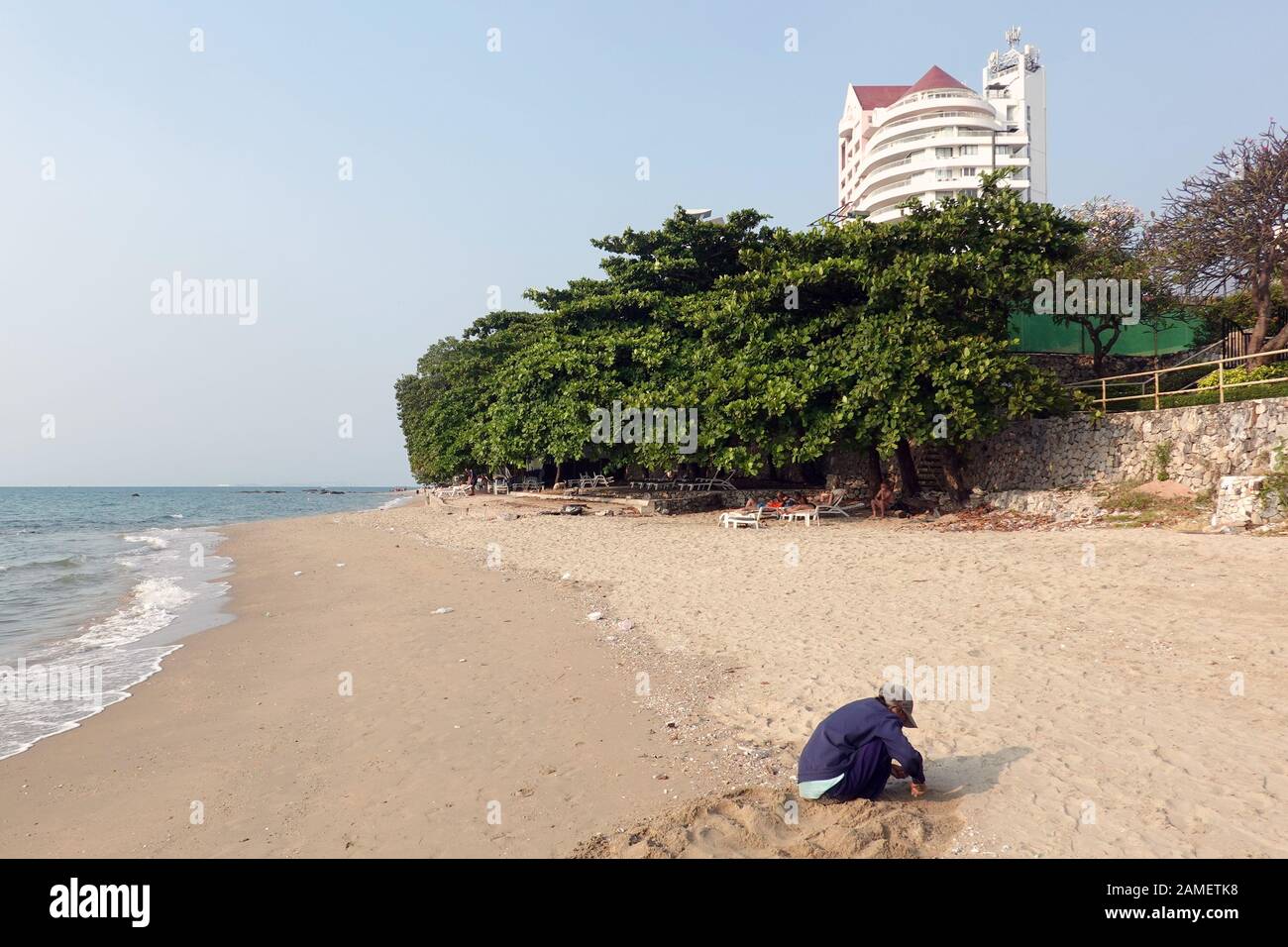 Pattaya, Thailand - 23. Dezember 2019: Ältere Frau sitzt am Strand und versucht, Muscheln zu finden. Ausländertouristen in Sonnenliegen im Hintergrund. Stockfoto