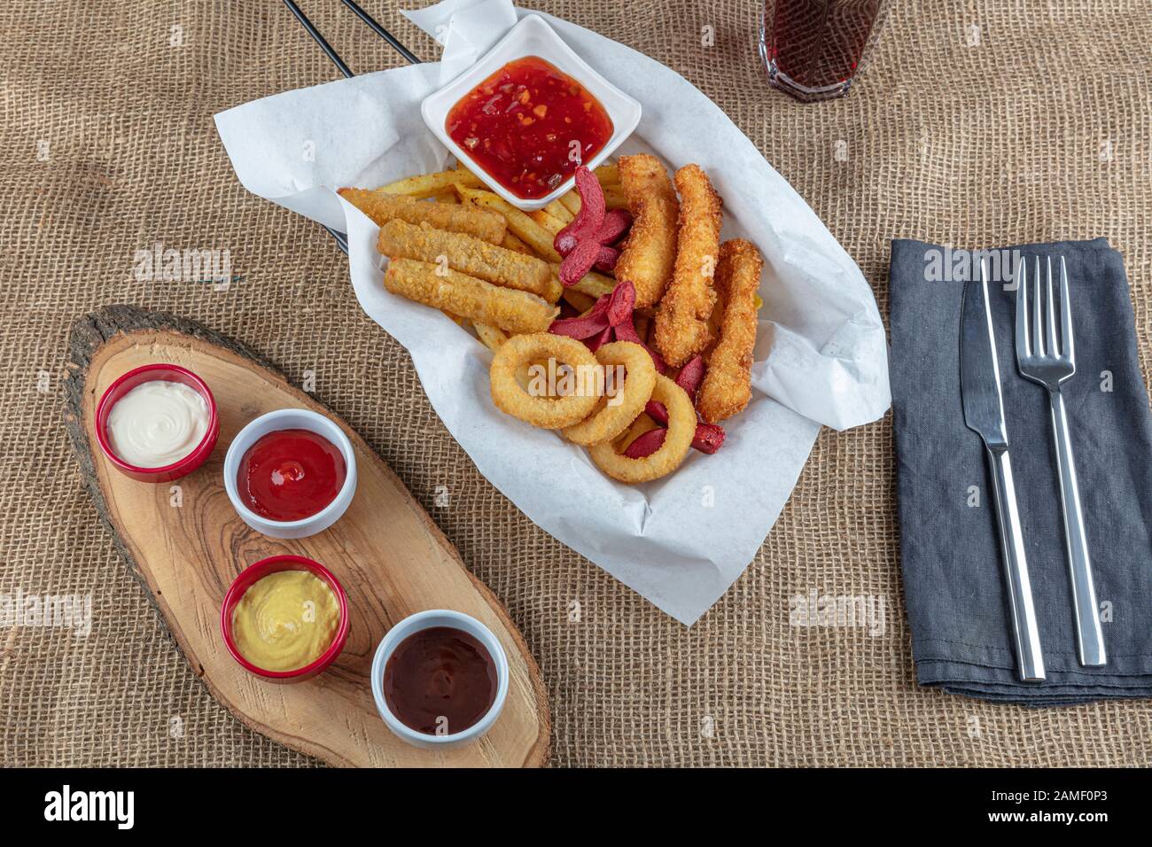 Imbissplatte - leckerer knolliger Zwiebelring, pommes frites, Wurst, gebratener Käse, heißer Imbiss. Spezielles Gewürz und Koks auf Leinengewebe. Stockfoto