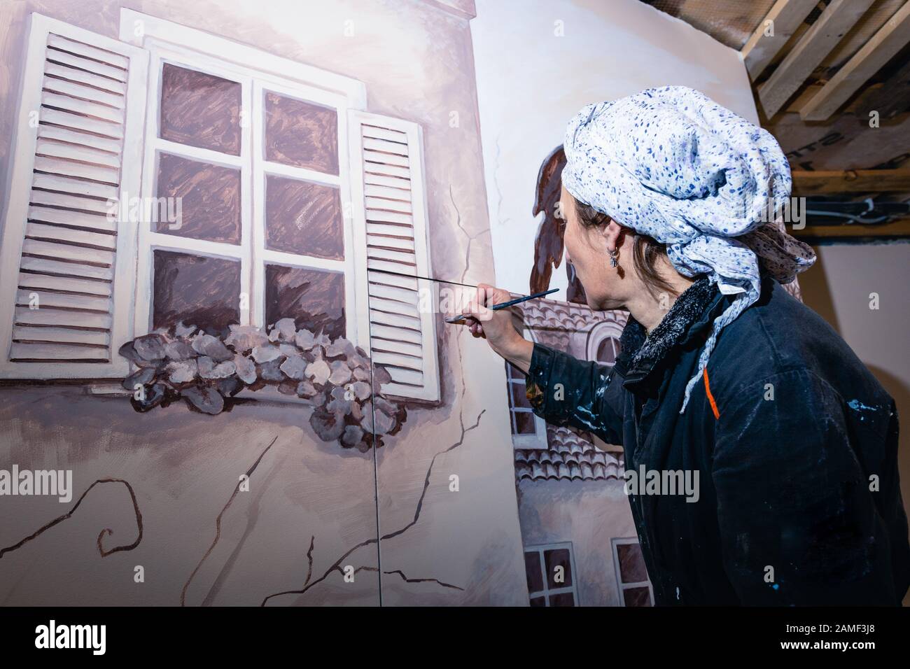 Frauenkünstlerin malt eine Wand in einem Wohnzimmer. Das Wandbild widmet sich den Eindrücken von Paris. Der Autor zeichnet ein Detail - Fensterläden. Rückansicht. Stockfoto