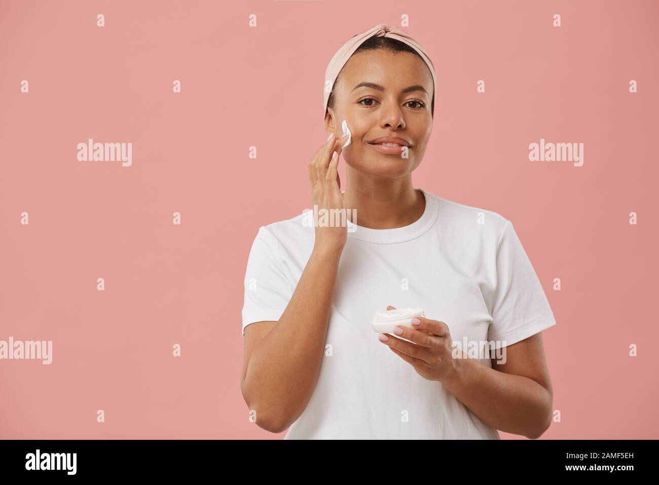 Taille hoch Portrait von Mixed-Race junge Frau mit Gesichtscreme und Lächeln auf der Kamera, während sie sich morgens vor pinkfarbenem Hintergrund während der Schönheitsroutine posiert Stockfoto