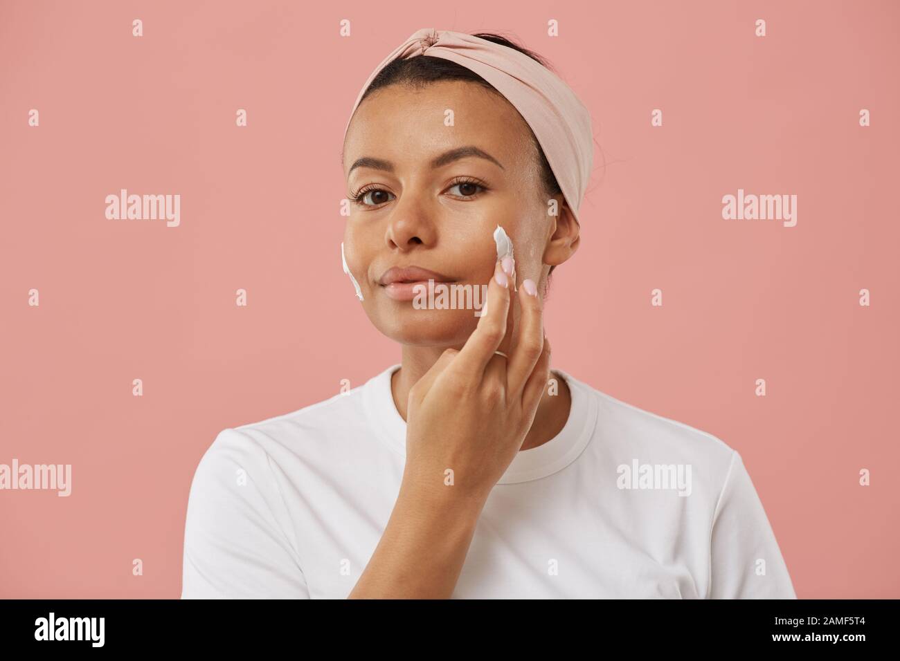 Porträt der schönen jungen Frau, die Gesichtscreme aufbringt und die Kamera betrachtet, während sie morgens bei der Schönheitsroutine vor rosafarbenem Hintergrund posiert Stockfoto