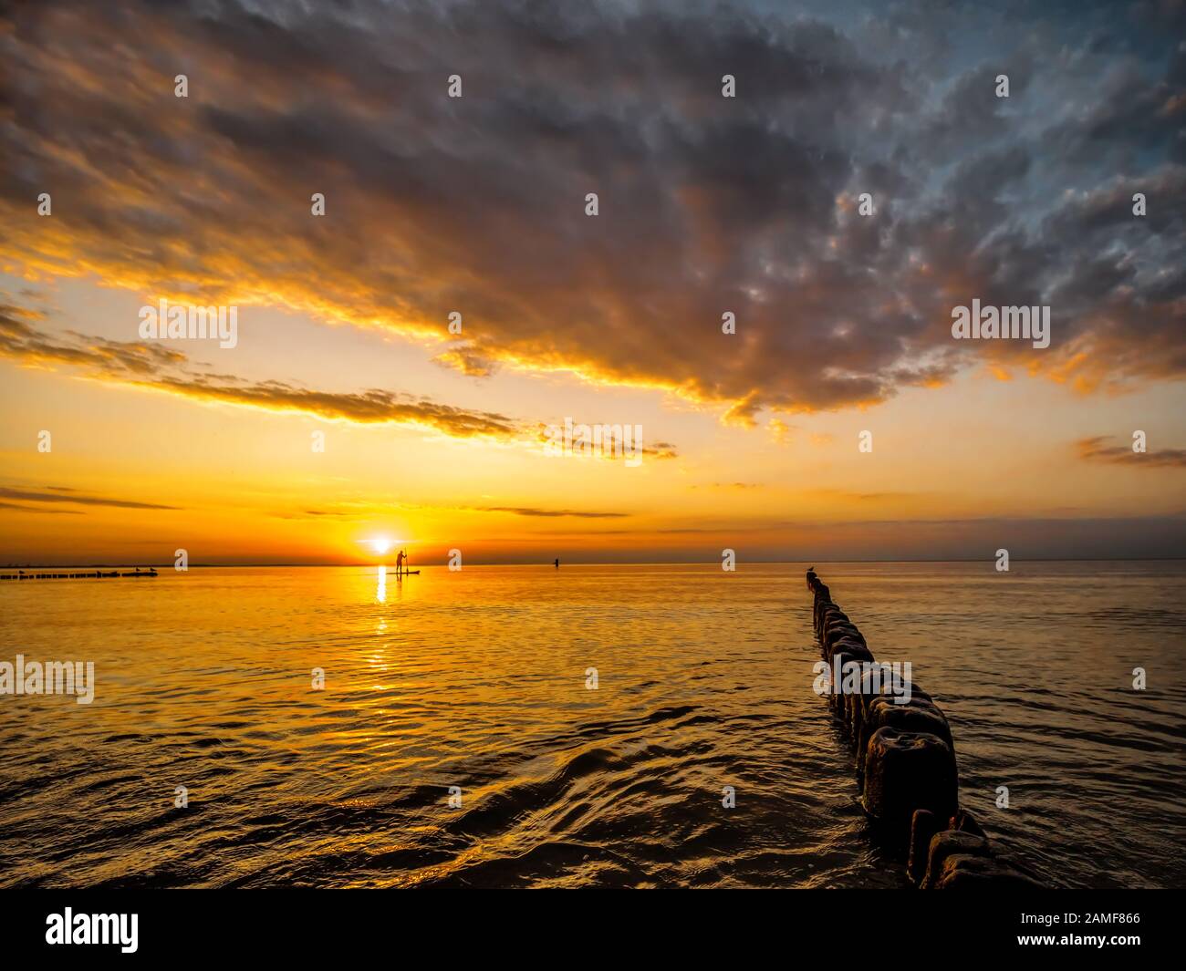 Silhouette einer Surfer surfen auf der Wasseroberfläche noch Meer gegen Sonnenuntergang und alte verwitterte Holz- Wellenbrecher im Vordergrund. Stockfoto