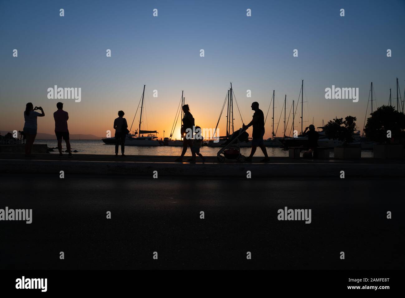 Silhouetten von Menschen, die den Sonnenaufgang und die Yacht-Masten bewundern, die von goldenem Stundenlicht in Naxos Town, Cyclades Group of Islands Greece, beleuchtet werden. Stockfoto