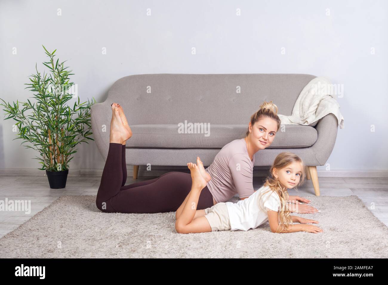 Attraktive junge Mutter, die Yoga-Sphinx-Pose mit kleiner Tochter praktiziert und Kamera betrachtet, glückliche Familie, die gemeinsam Turnen macht, fitne Stockfoto