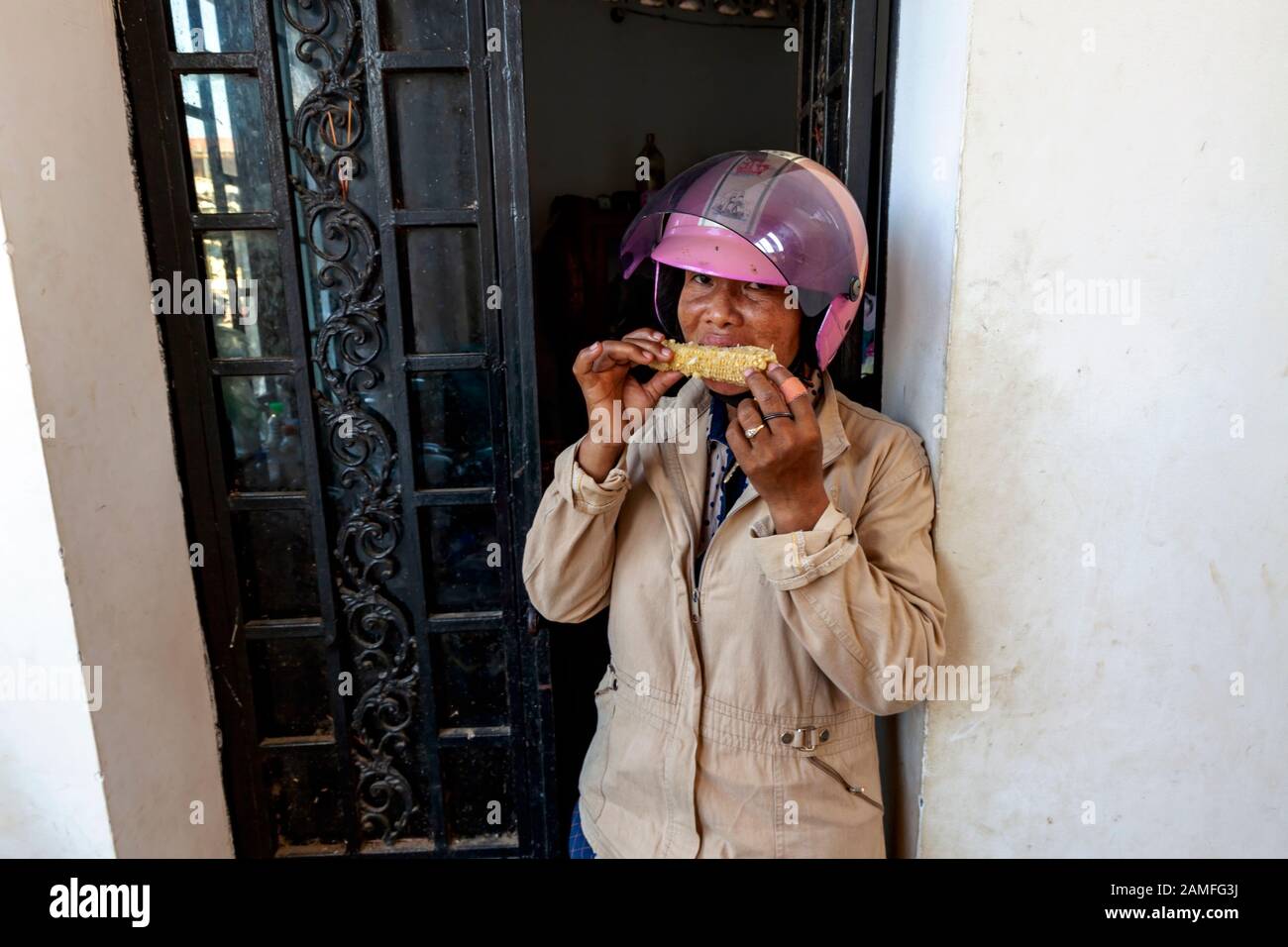 Eine asiatische Frau mittleren Alters, die einen rosafarbenen Motorradhelm auf dem Kopf trägt, isst ein Stück Mais im städtischen Kampong Cham, Kambodscha. Stockfoto
