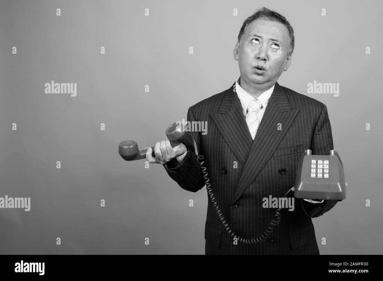 Studio von reifen asiatischen Geschäftsleuten mit altem Telefon vor grauem Hintergrund in Schwarzweiß gedreht Stockfoto