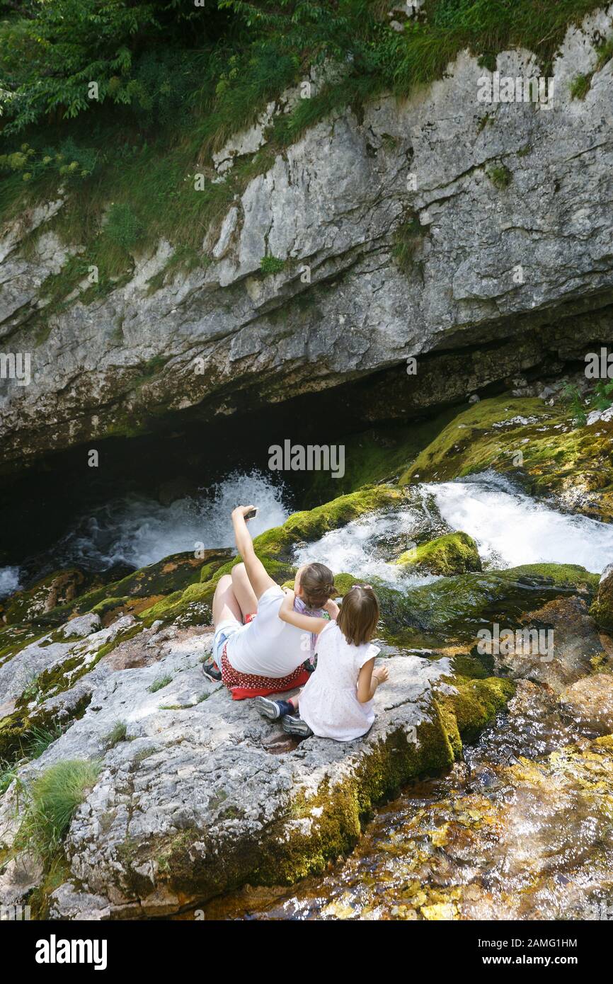 Mutter mit Kindern, die ein selfie nehmen, umarmen und auf einem Felsen an einem Bergbach auf einem Familienausflug sitzen. Outdoor-Lifestyle, natürliche Erziehung, Kindheit Stockfoto
