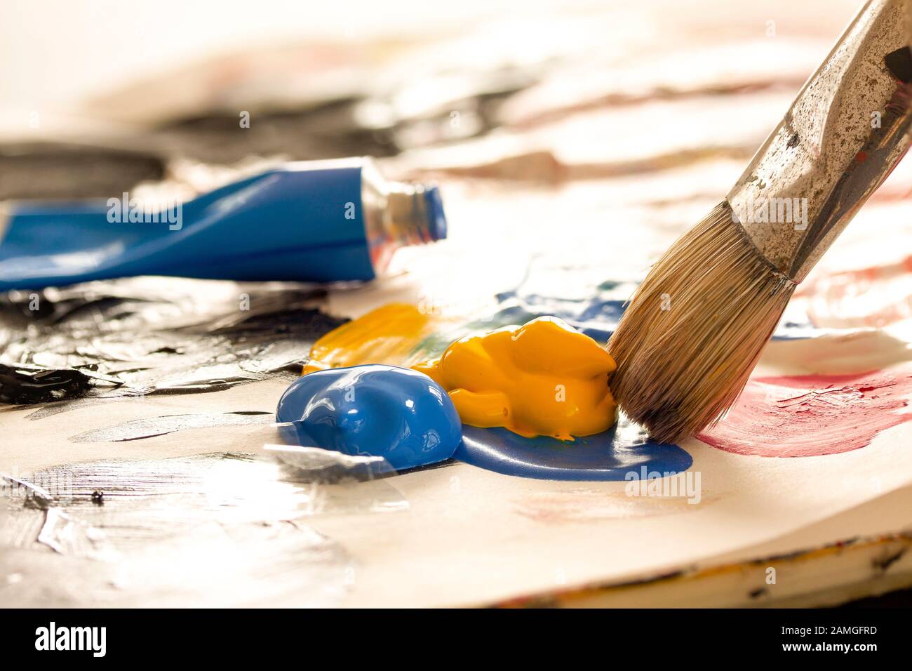 Künstlerbürste mit Lack aus Malröhrchen, die auf einer Palette vermischen können. Blaue Farbröhre im Hintergrund. Nahaufnahme mit Hintergrundbeleuchtung und selektivem Fokus. Stockfoto