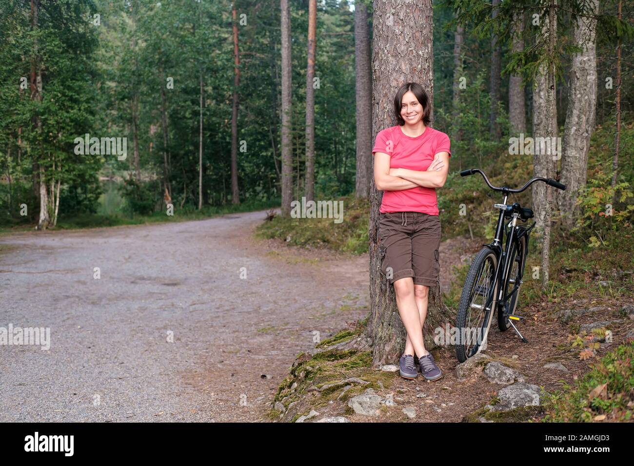 Lächelnde Frau lehnt sich an einen Baum, ruht und genießt die Landschaft, neben ihr befindet sich ein altes Fahrrad, in einem schönen Wald. Stockfoto