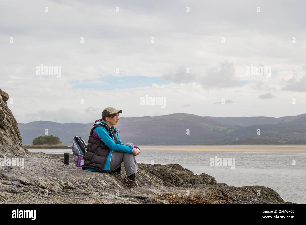 Isolierte junge Frau in Wanderausrüstung & Sportmütze, sitzend auf Felsen an der Küste mit Blick auf das Meer, tief in Gedanken, Wales Großbritannien. Aufladen der Akkus. Stockfoto