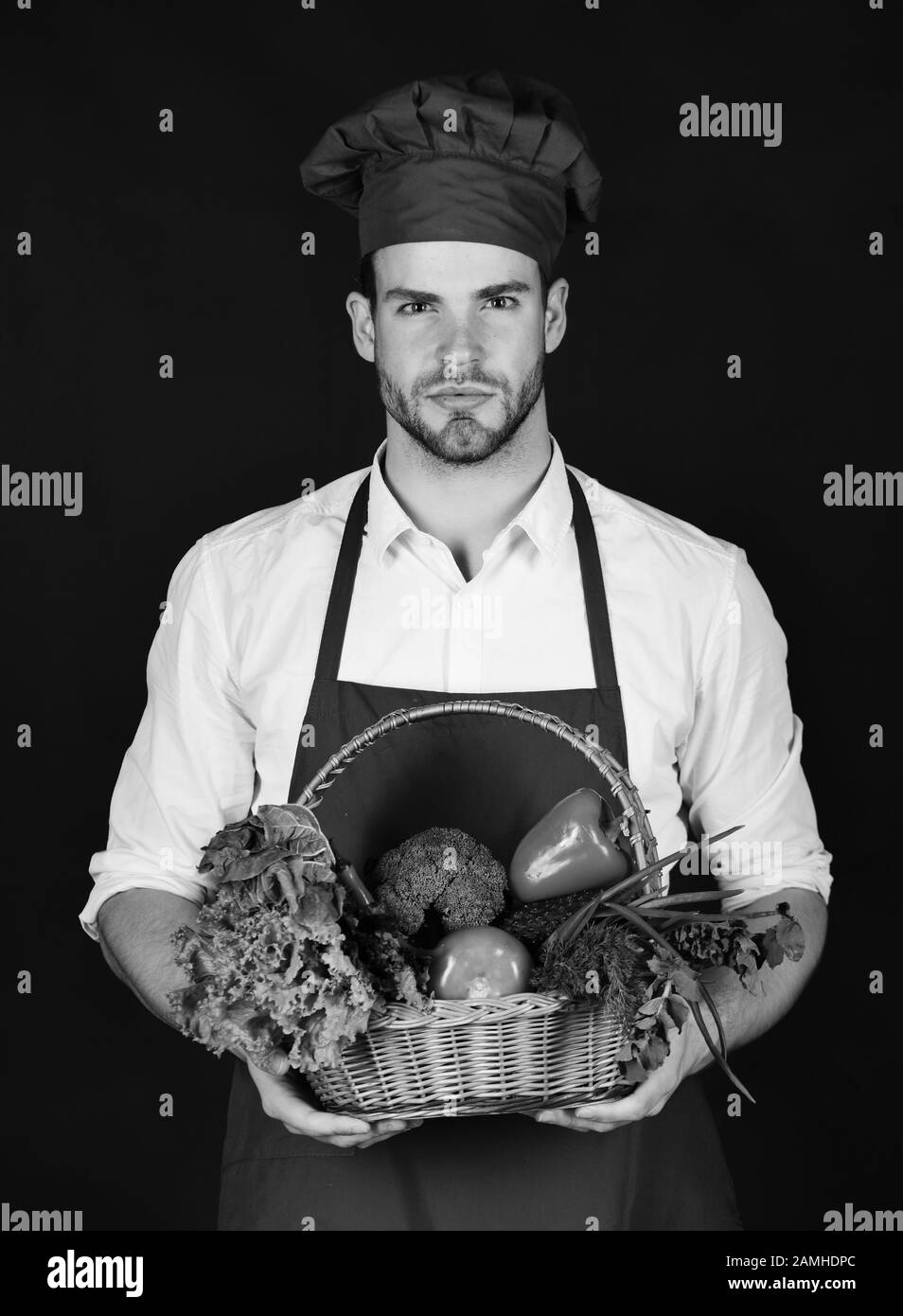 Küchenchef in burgunder Uniform hält Korbkorb mit Gemüse. Mann mit Bart und Korb mit Gemüse auf schwarzem Hintergrund. Kochen mit ernsthaftem Gesicht hält frische Ernte. Vegetarisches Diät-Konzept. Stockfoto