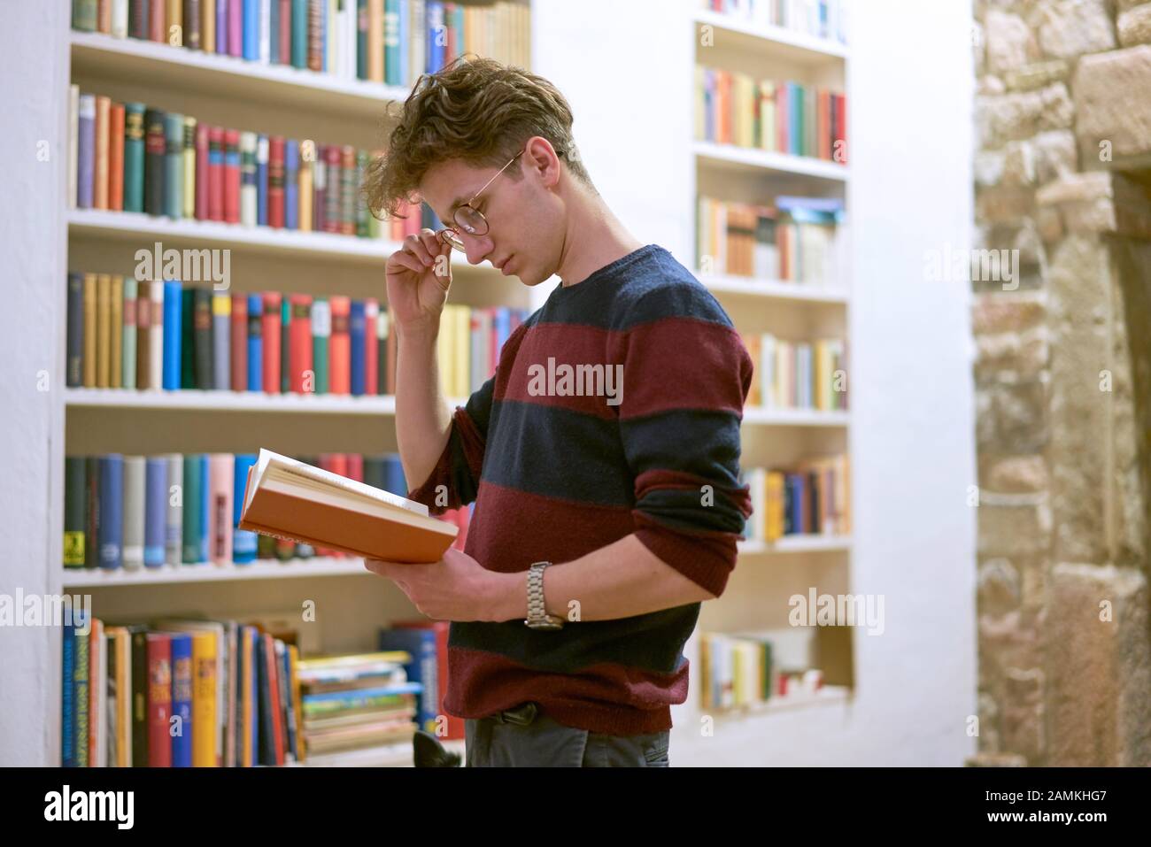 Geballte junge Studentin mit Brille, die ein Buch in der Bibliothek neben dem Bücherregal liest. Steinwand im Hintergrund. Stockfoto