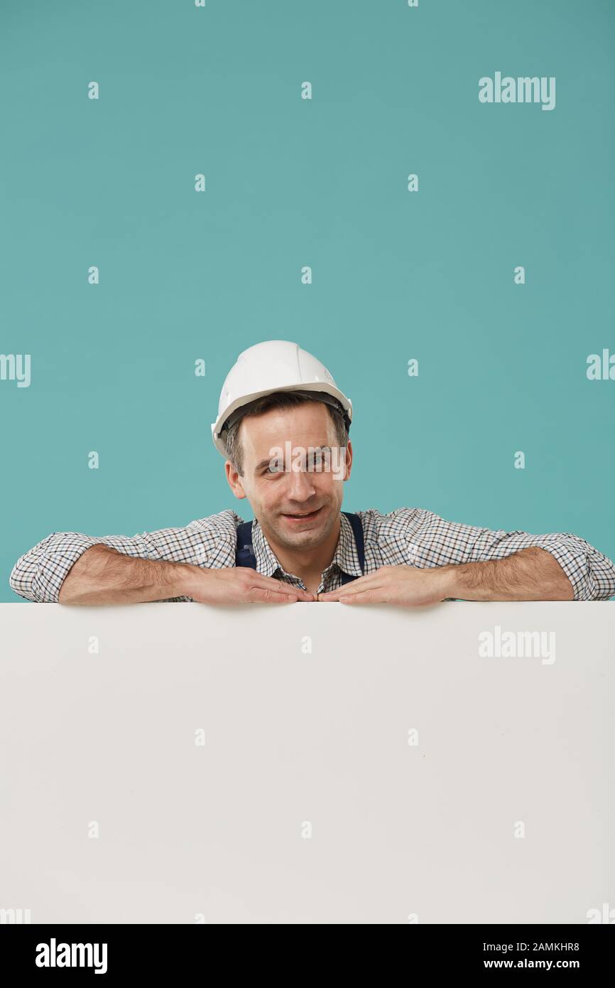 Porträt eines lächelnden Mitarbeiters, der einen Hardhat trägt, der sich vor blauem Hintergrund über ein leeres weißes Schild stützt, Platz für Kopien Stockfoto