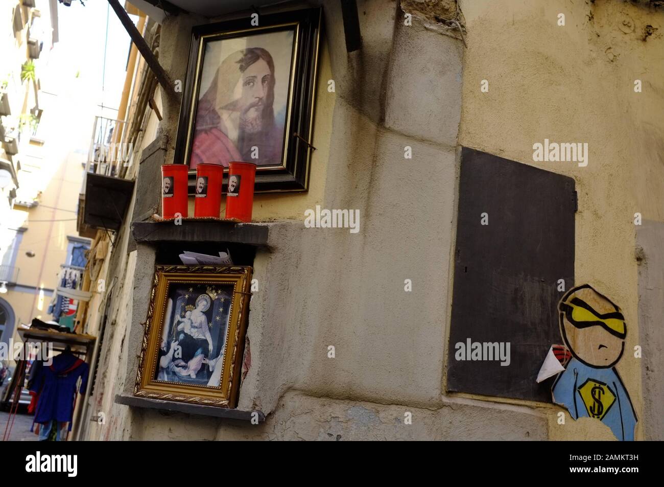 Ein eingerahmtes Jesusgemälde, davor drei Kerzen und eine Darstellung der Jungfrau Maria an der Ecke eines Hauses in der Altstadt von Neapel. Daneben moderne Kunst, ein Graffiti - Kopf mit verbundenen Augen und Dollarzeichen auf der Brust. [Automatisierte Übersetzung] Stockfoto