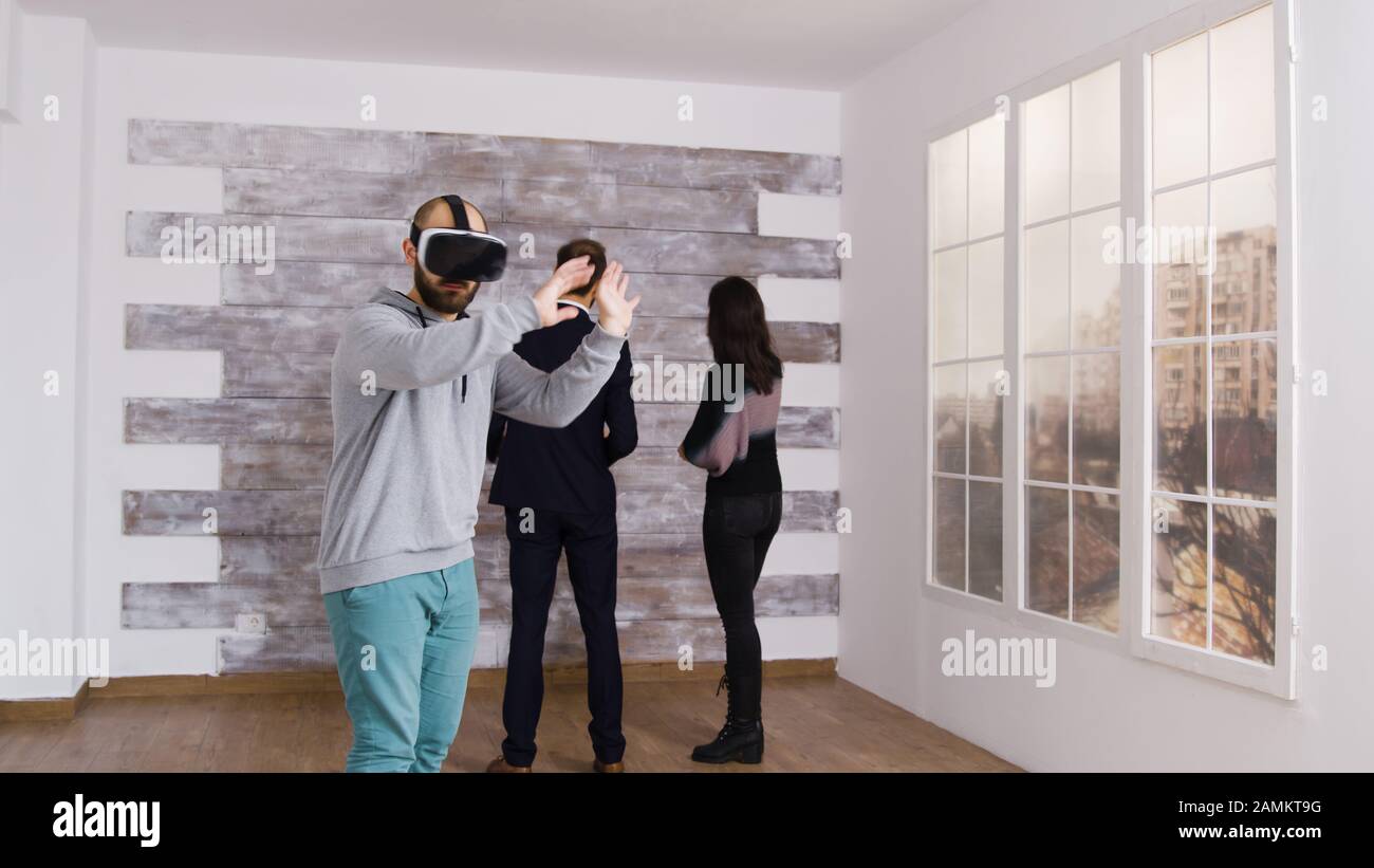 Kaukasischer Mann mit Virtual-Reality-Headset in leerer Wohnung, während eine Frau mit Immobilienmakler spricht. Stockfoto