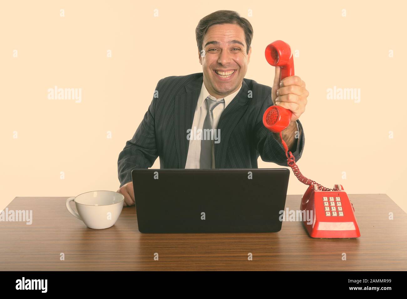 Glücklicher persischer Geschäftsmann, der bei der Arbeit ein altes Telefon mit Laptop telefonieren lässt Stockfoto