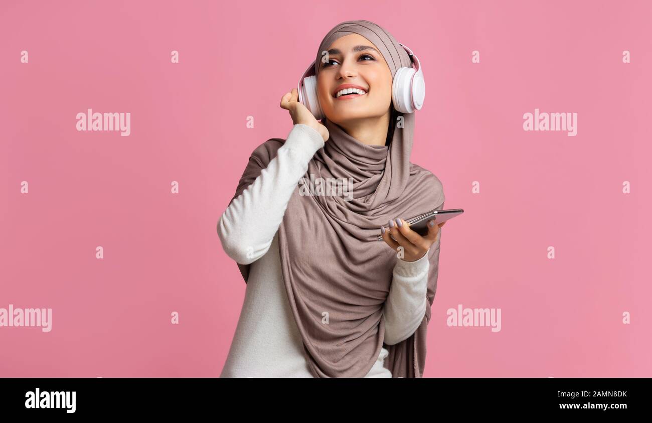 Positive muslimische Frau in Hijab und Kopfhörer, die Musik auf dem Smartphone hören Stockfoto