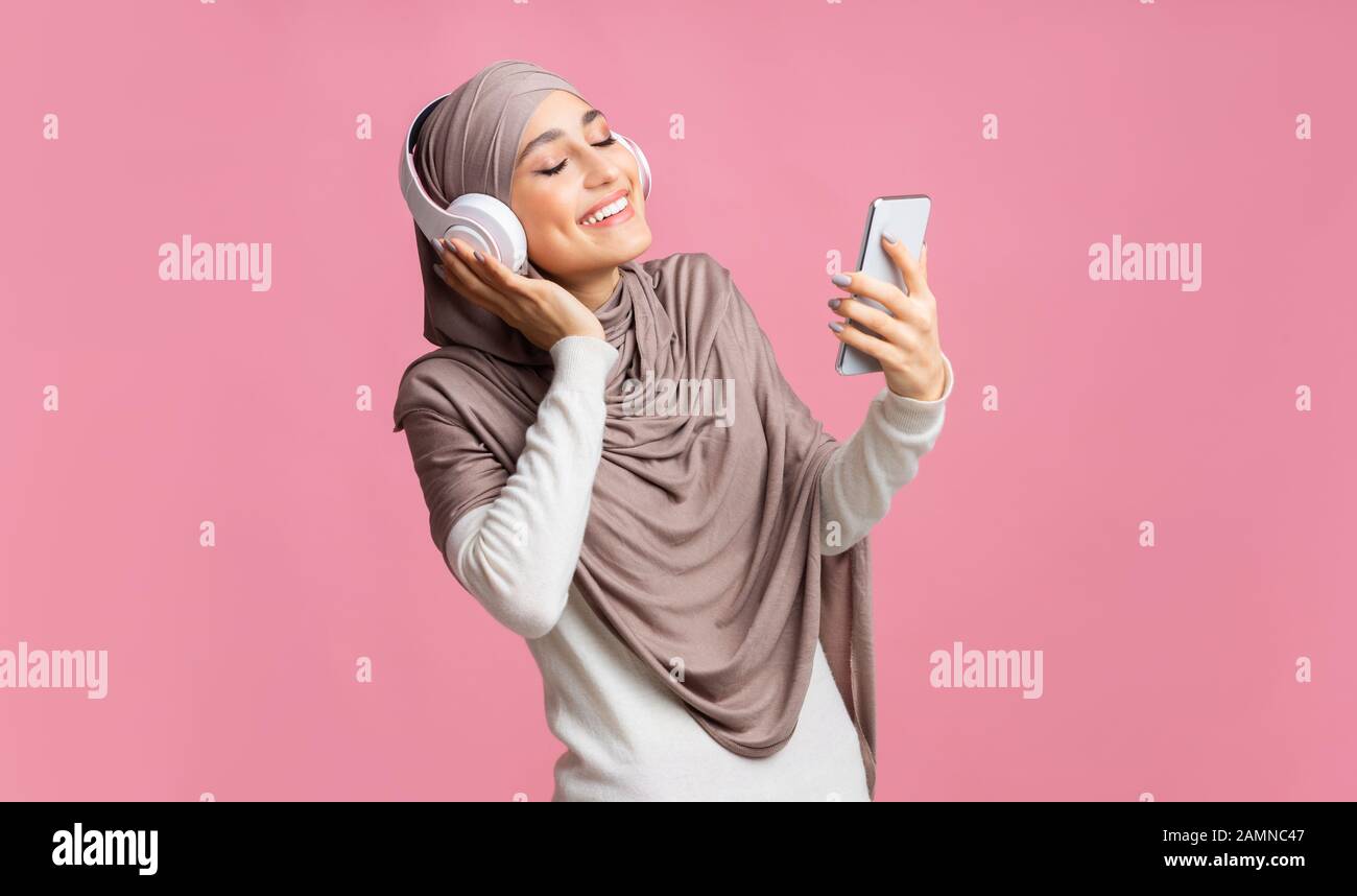 Fröhliches muslimisches Mädchen in kabellosen Kopfhörern, das online Musik auf dem Smartphone hört Stockfoto