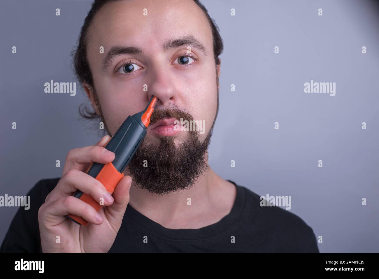 Portrait eines jungen bärtigen Kerls, verwendet einen Trimmer, um Nasenhaare zu schneiden. Gesundheit und Schönheit der Männer. Auf grauem Hintergrund: Junger Kerl schneidet Nasenhaar mit Trimme Stockfoto