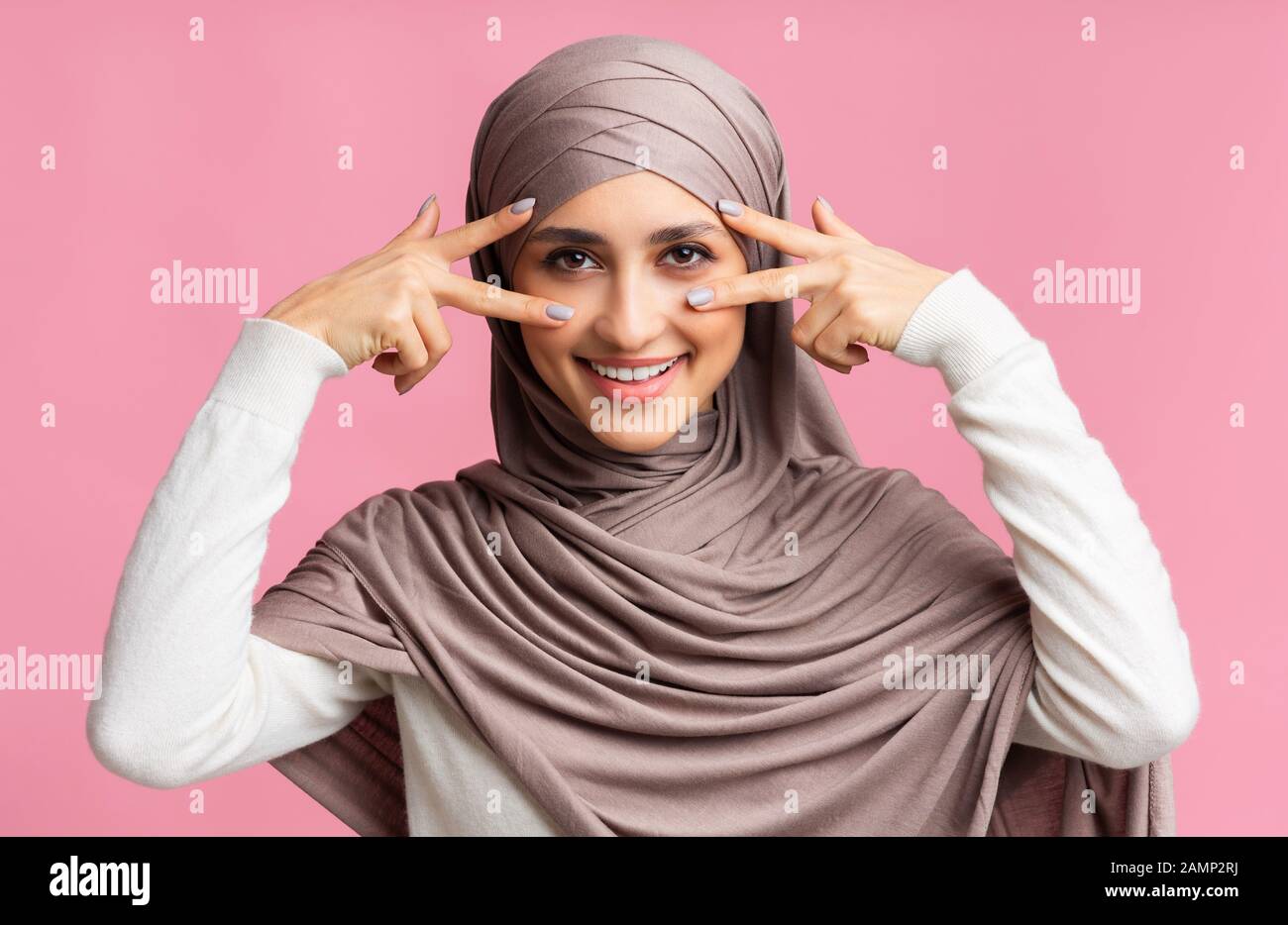 Verspielte muslimische Frau im Hijab, die V-Zeichensymbole in Augennähe zeigt Stockfoto