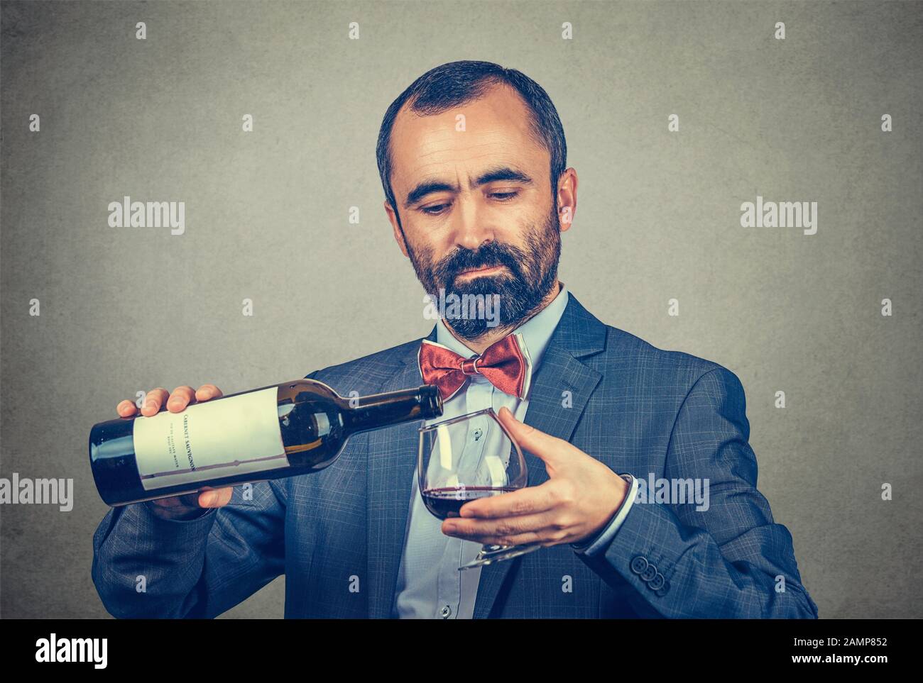 Ein Mann gießt etwas Wein aus einer Flasche mit Blick auf die Weinfarbe in ein Glas. Gemischtes Rennmodell auf grauem Hintergrund mit Kopierbereich isoliert. Stockfoto