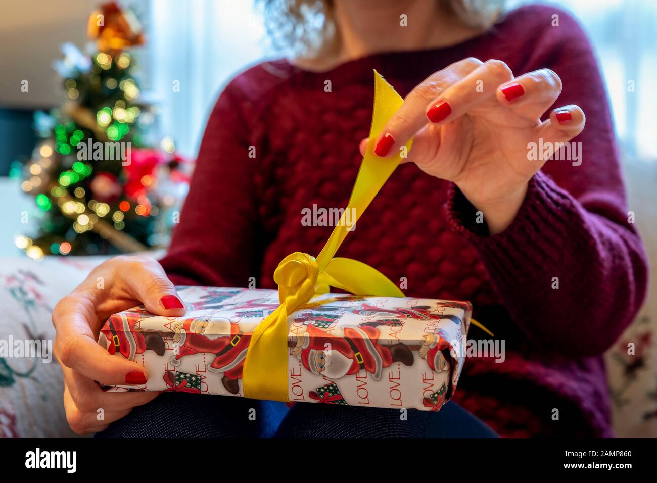 Blonde Frau enthüllt ein Weihnachtsgeschenk mit dem Weihnachtsbaum im Hintergrund, wobei die Lichter aufleuchten Stockfoto