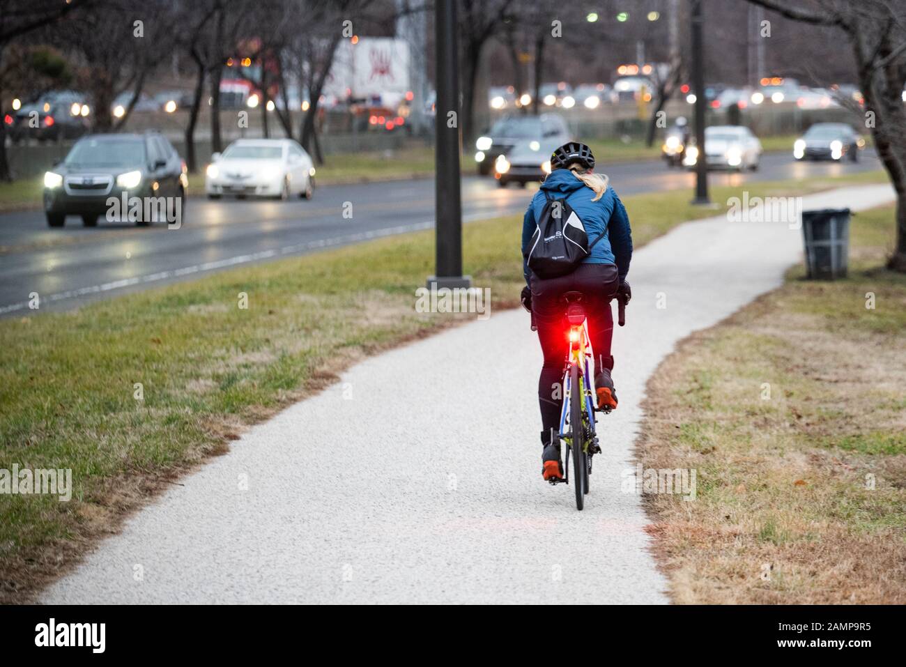 Ein Radfahrer, der am frühen Morgen auf einem Radweg unterwegs ist, mit einer roten Heckleuchte und Fahrzeugverkehr im Hintergrund. Stockfoto