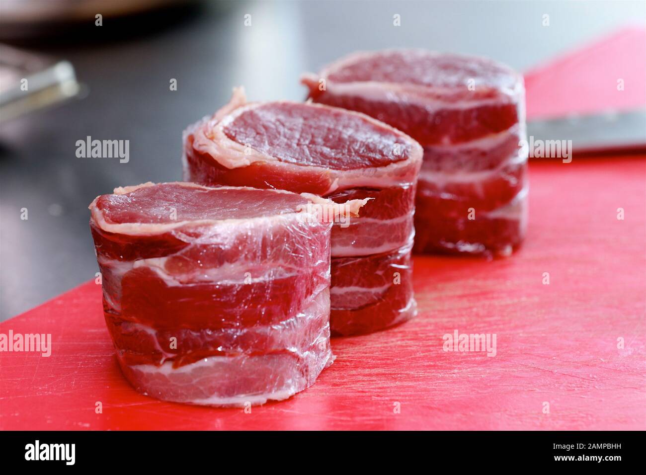Rohes Filet von Hereford Irish Beef Filet Steak in parmaschinken auf einem roten Hackbrett mit Messer im Hintergrund eingewickelt. Stockfoto