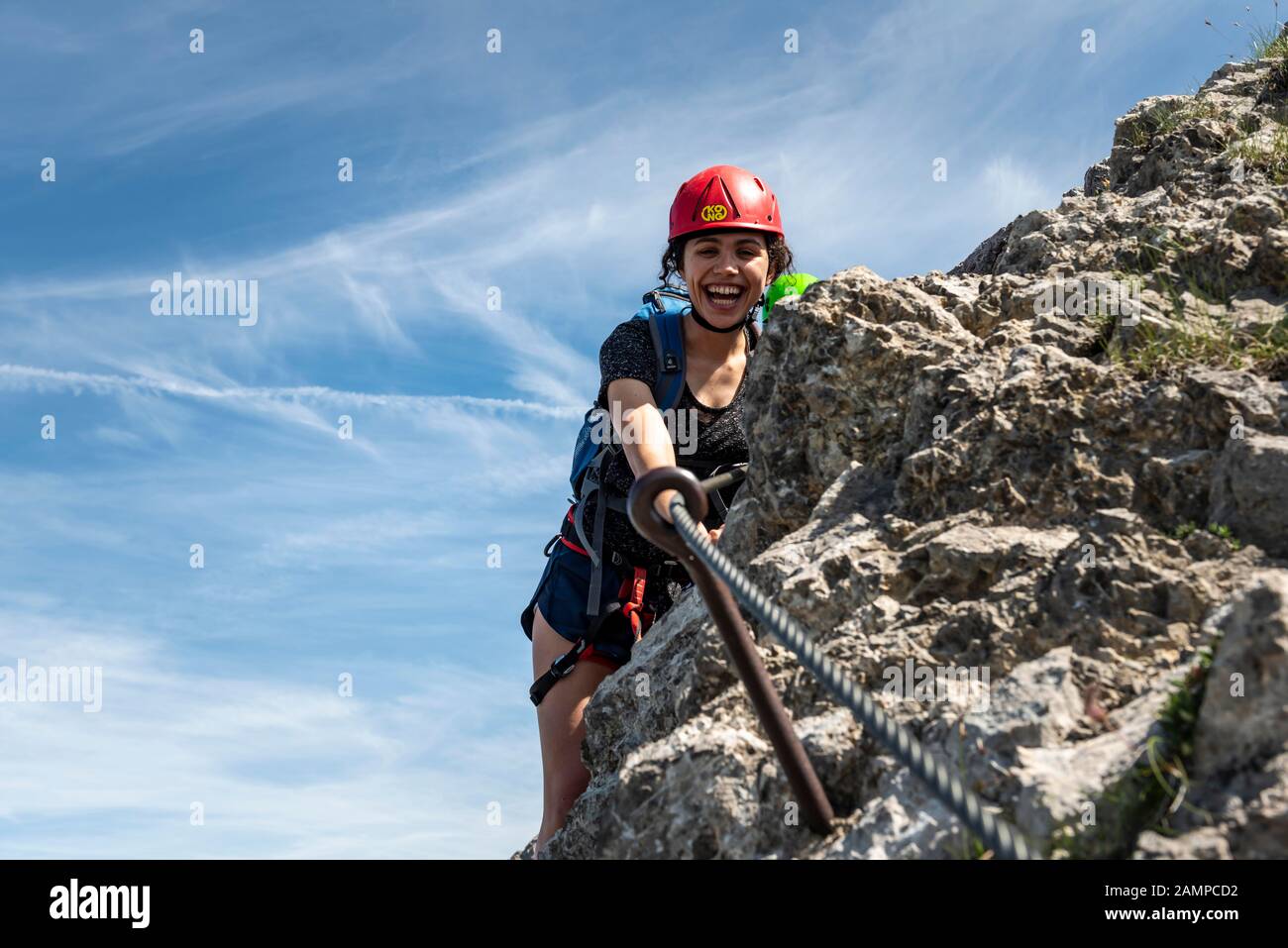 Lachende junge Frau mit Kletterhelm und auf dem mit Seil gesicherten Felsen, weibliche Kletterin auf einem Klettersteig, Mittenwald über Ferrata, Karwendel Stockfoto