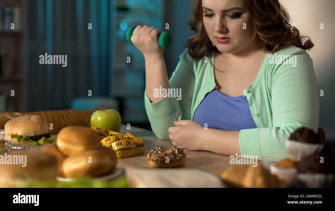 Fette Dame, die mit Hanteln trainiert und süße Donut, nährende Willenskraft betrachtet Stockfoto
