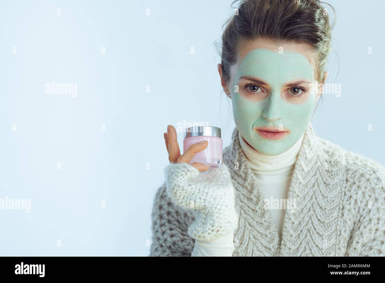 Porträt der modernen Hausfrau in Rollkragen-Pullover und Strickjacke mit grüner Gesichtsmaske, die Kosmetikbecher isoliert im Winter hellblau hält. Stockfoto
