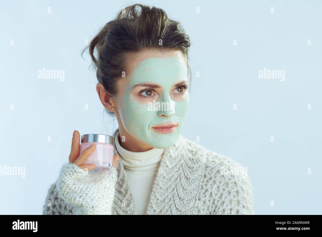 Moderne Frau mit Rollkragen-Pullover und Strickjacke mit grüner Gesichtsmaske, die einen Kosmetikbecher mit Blick auf den Kopierbereich vor dem hellblauen Hintergrund im Winter hält. Stockfoto