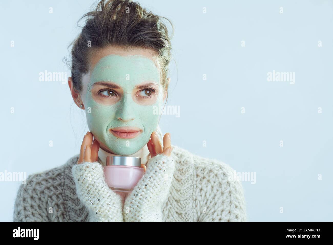 Elegante Frau in Rollkragen-Pullover und Strickjacke mit grüner Gesichtsmaske, die Kosmetikbecher beiseite hält, isoliert im Winter hellblau. Stockfoto