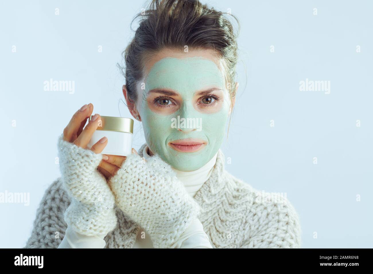 Moderne Frau im mittleren Alter mit Rollkragen-Pullover und Strickjacke mit grüner Gesichtsmaske, die einen kosmetischen Mixbecher auf winterhellblauem Hintergrund zeigt. Stockfoto