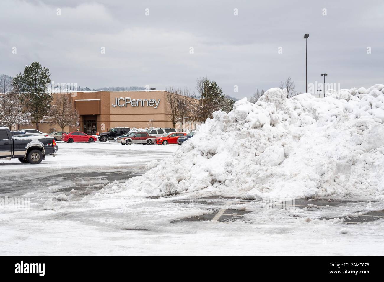 Ein massiver Haufen gepflügten Schnees nach einem Wintersturm sitzt auf einem Parkplatz eines Einkaufszentrums mit Kaufhaus im Blick dahinter. Stockfoto