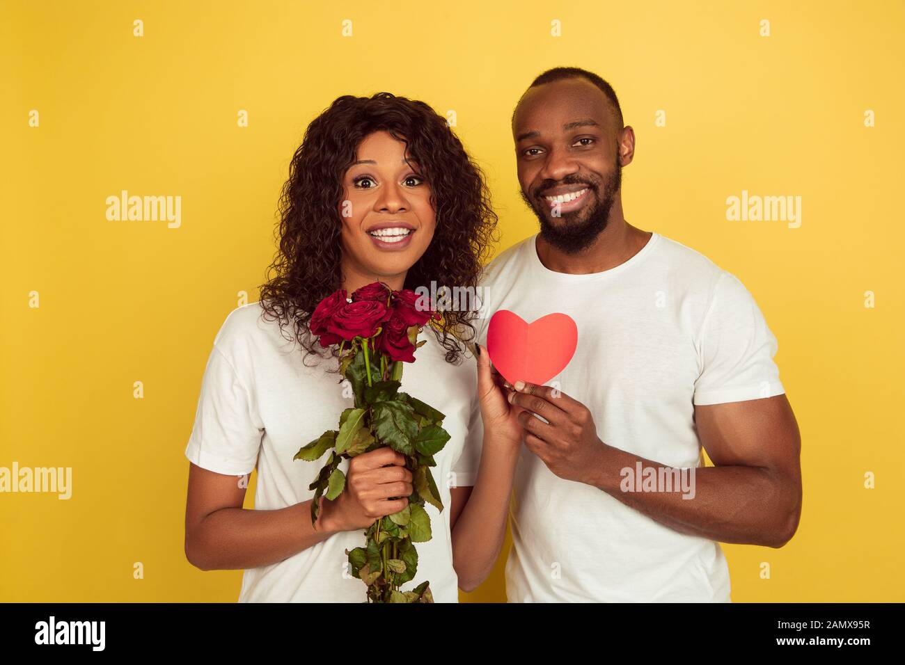 Blumen und Herz. Valentinstag gefeiert, glückliches afro-amerikanisches Paar isoliert auf gelbem Studiohintergrund. Konzept menschlicher Emotionen, Gesichtsausdruck, Liebe, Beziehungen, romantische Feiertage. Stockfoto