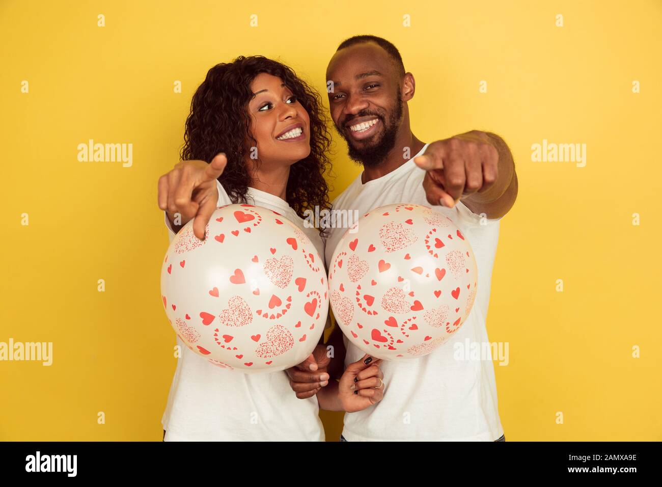 Zeigen mit Luftballons. Valentinstag gefeiert, glückliches afro-amerikanisches Paar isoliert auf gelbem Hintergrund. Konzept menschlicher Emotionen, Gesichtsausdruck, Liebe, Beziehungen, romantische Feiertage. Stockfoto