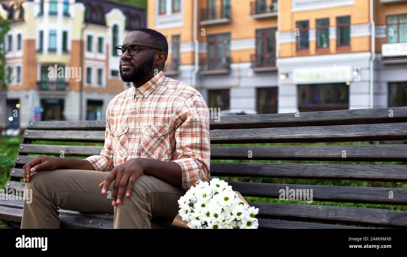 Trauriger Mann wartet nervös auf Mädchen, sitzende Parkbank mit Blumen, blindes Datum Stockfoto