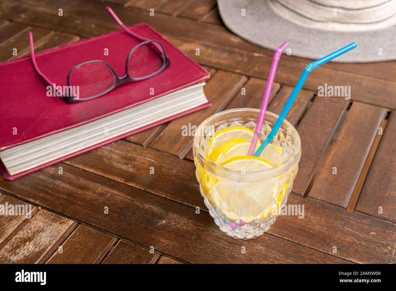 Kaltes Getränk mit Eis und Zitronenscheiben, Buch und Hut auf Holztisch. Freizeit oder Lesekonzept zur Sommerzeit Stockfoto