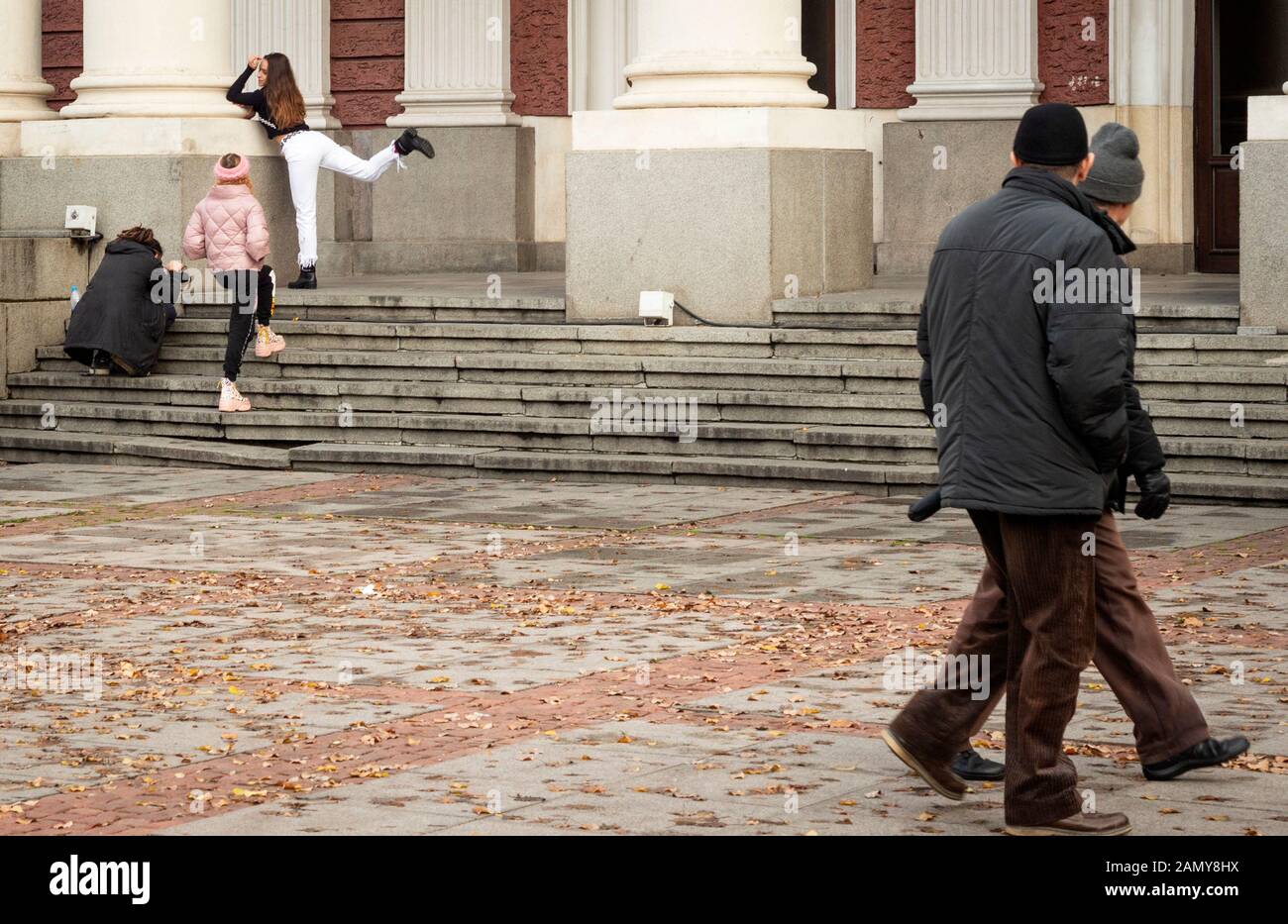 Generationslücke zeigt sich als zwei erfahrene Passanten, die eine Gruppe von Teenagern beobachten, die in Sofia, Bulgarien, Osteuropa, dem Balkan und der EU posieren und Fotos machen Stockfoto