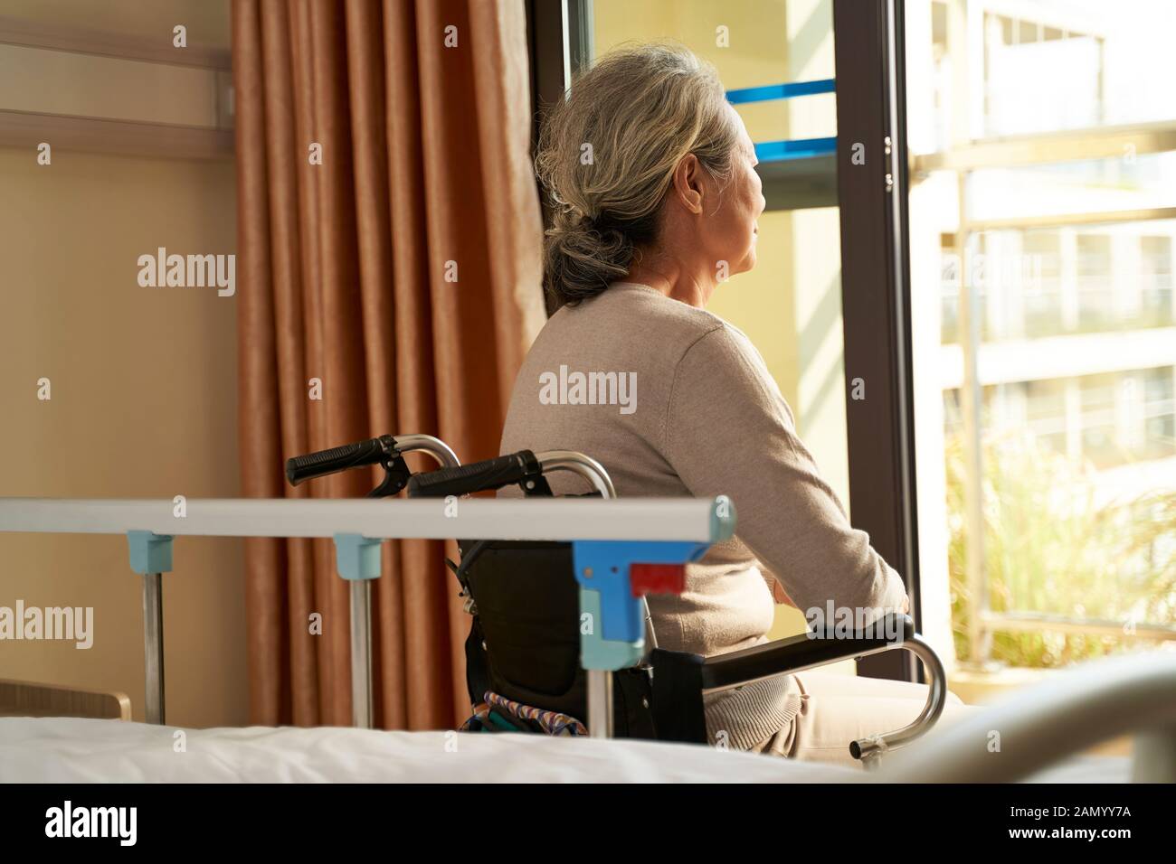 Rückansicht der älteren asiatischen Frau, die im Rollstuhl sitzt, in der Hausstation oder auf der Krankenstation, die aus dem Fenster blickt Stockfoto