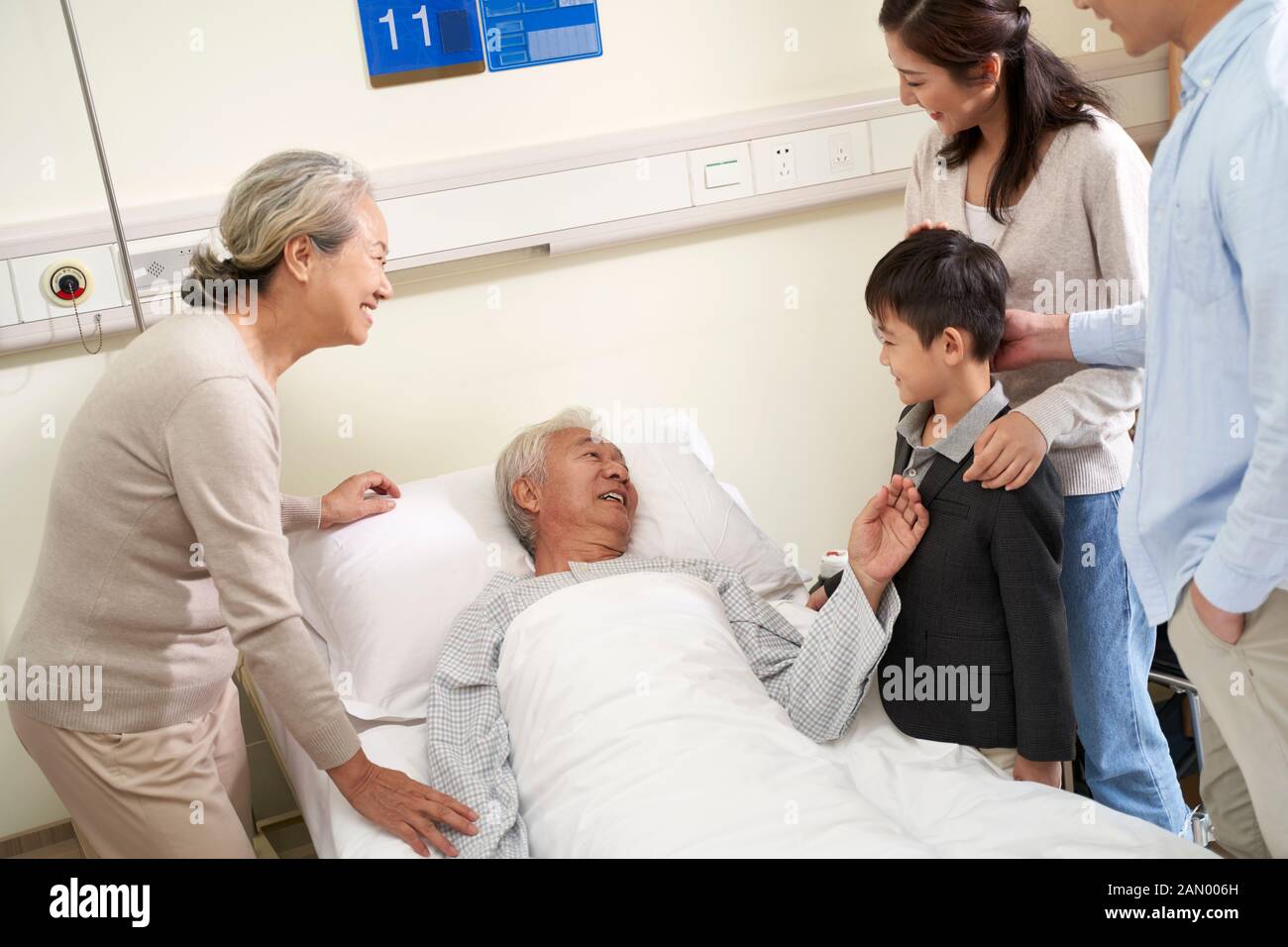 Glückliche asiatische Familie der drei Generationen, die mit dem hospitalisierten Großvater am Krankenbett spricht Stockfoto