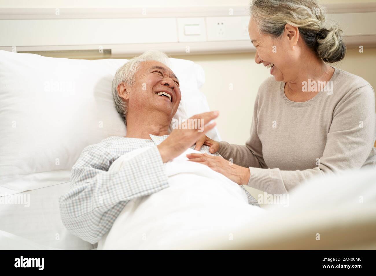 Liebevolle und fürsorgliche ältere asiatische Frau, die am Krankenbett einen Ehemann besucht und mit einem hospitalisierten Mann spricht Stockfoto
